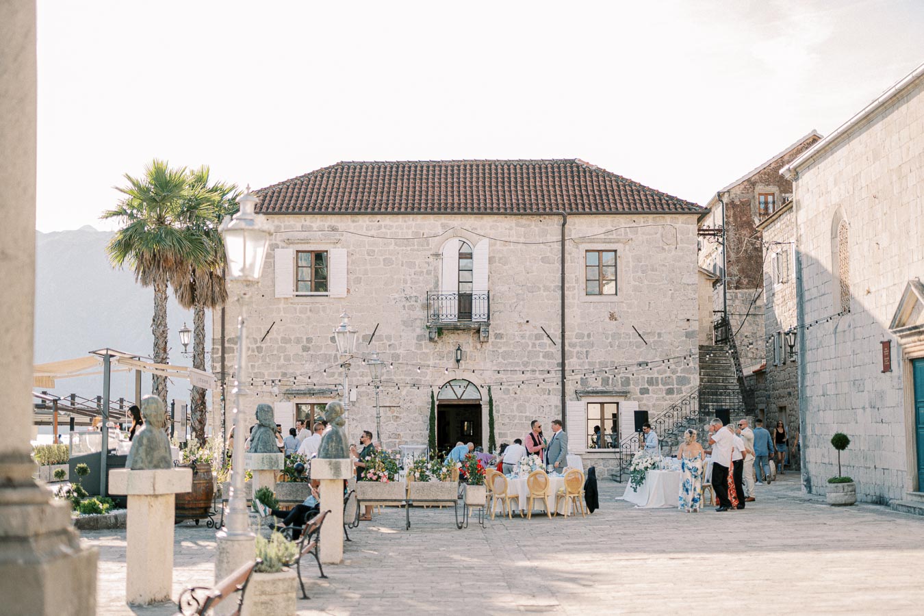 Ancient stone building with outdoor gathering in Mediterranean town square featuring palm trees, statues, and decorative string lights.