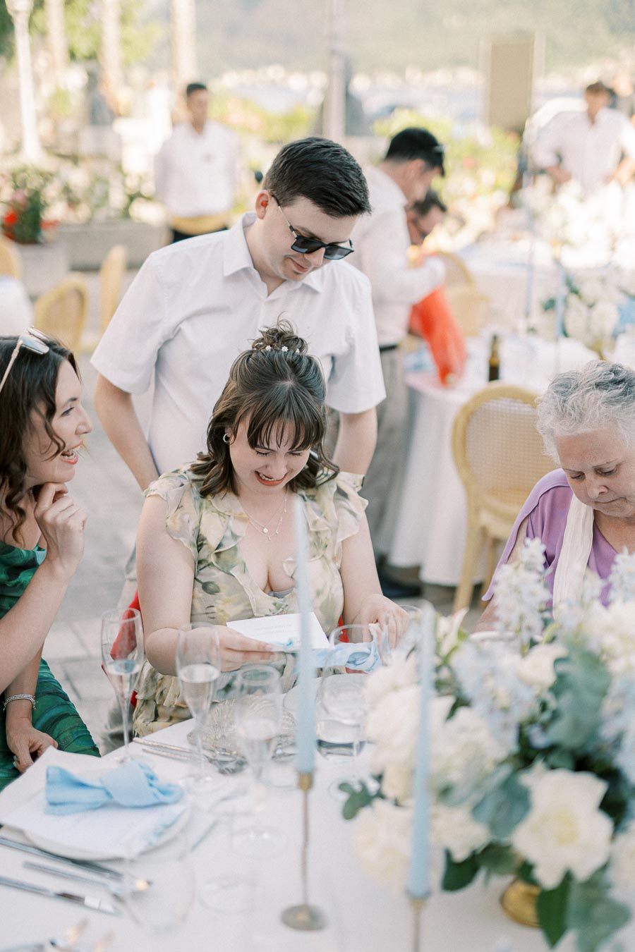 A group of people at an outdoor event, gathered around a table adorned with white and blue flowers and candles. A woman reads a card, smiling, while others look on, creating a cheerful atmosphere.