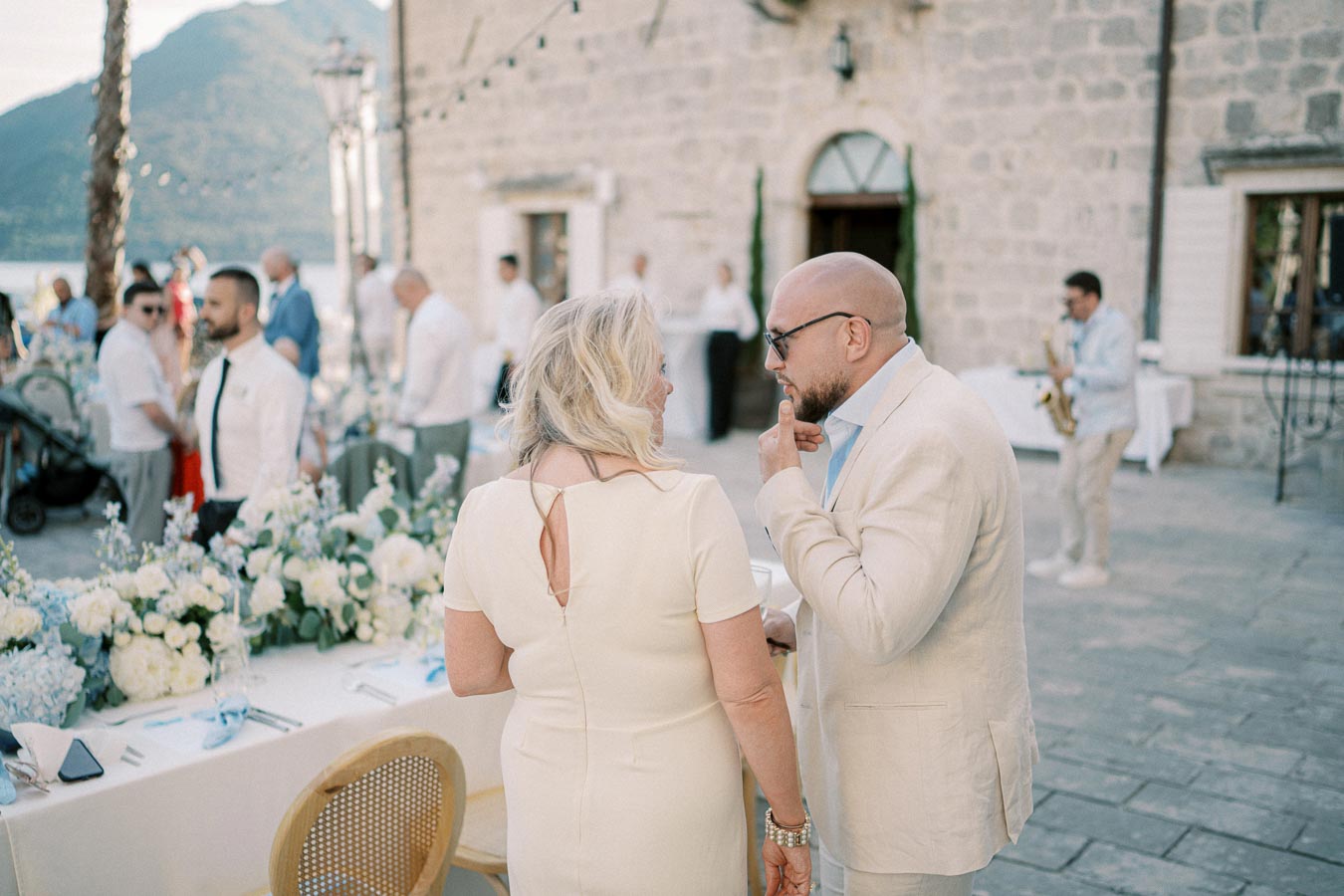 Elegant outdoor wedding reception with guests in formal attire engaged in conversation. The setting features a beautifully decorated table with floral arrangements and a historical stone building in the background. A musician plays the saxophone, creating a festive atmosphere.