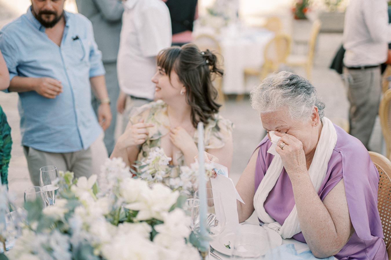 Elderly woman wiping tears at a wedding reception, surrounded by guests and a table decorated with flowers.