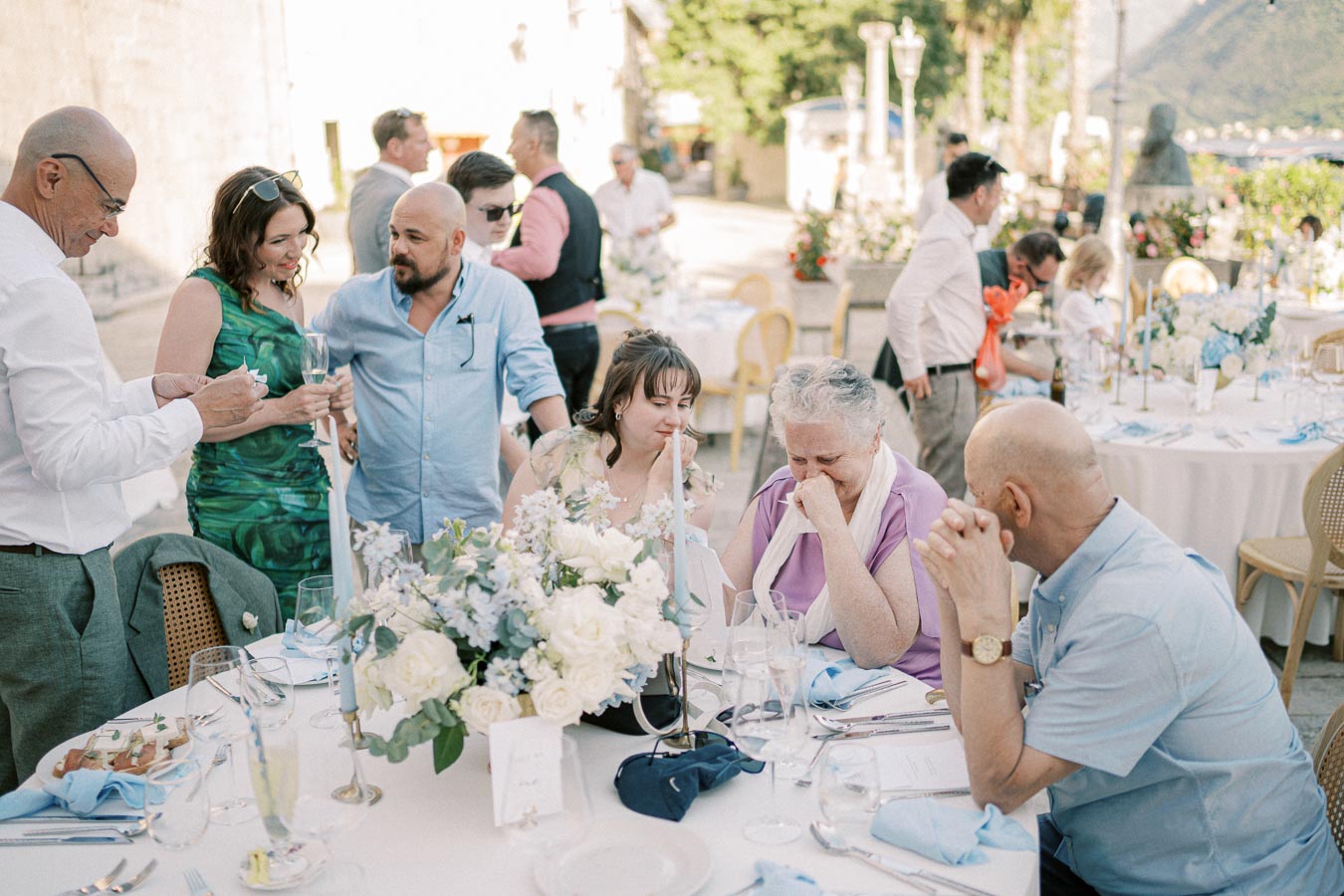 Outdoor wedding reception with guests mingling around a beautifully decorated table adorned with white and blue floral arrangements. People are engaged in conversation and enjoying a sunny day in a scenic setting, providing a relaxed and festive atmosphere.