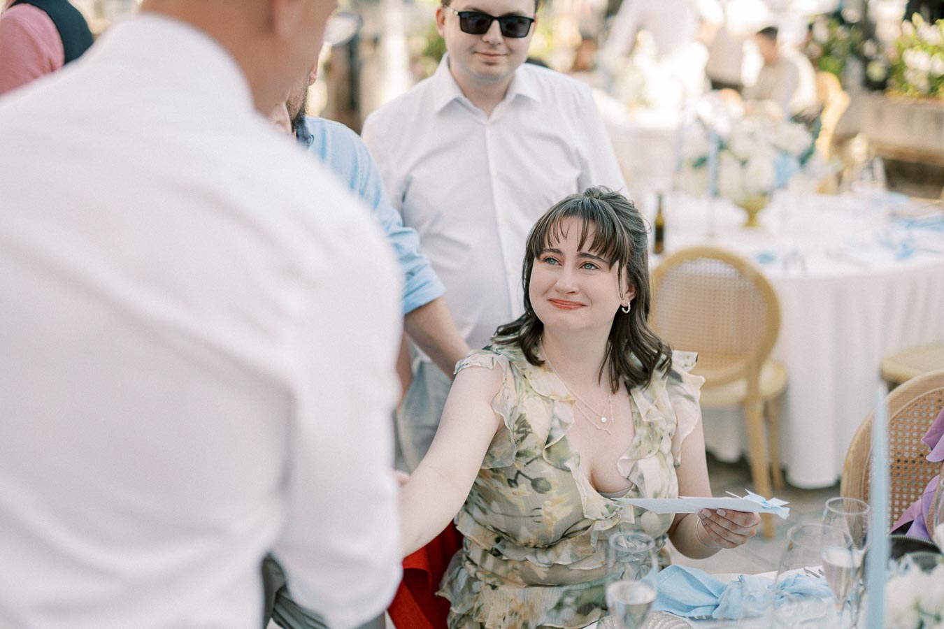 A woman in a floral dress interacts with guests at an outdoor event, seated at a table with white tablecloths and blue decorations, while handing a paper to a man in a white shirt.
