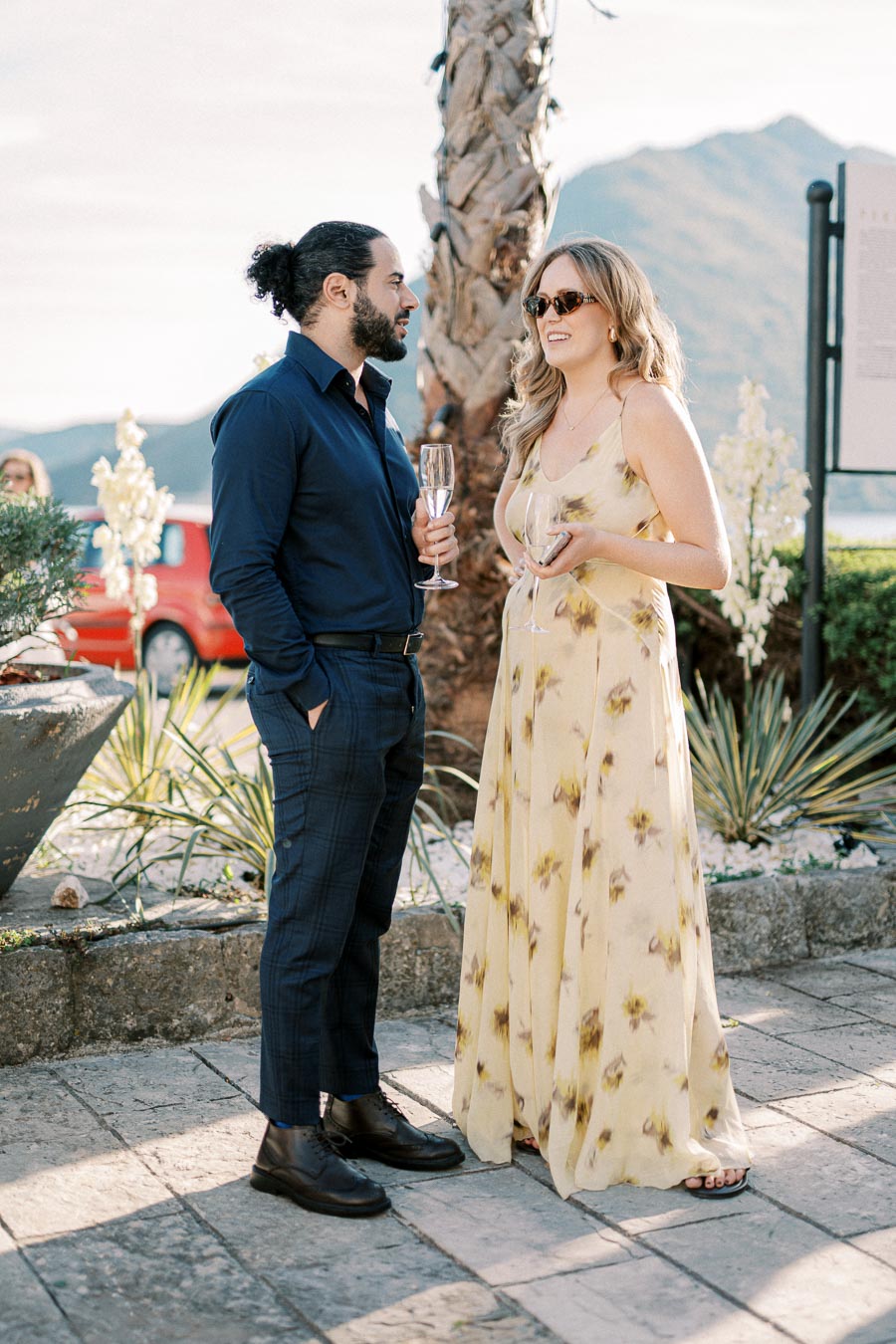 Two people enjoying a conversation outdoors at a social event, dressed elegantly with the man in a navy shirt and checkered pants, and the woman in a floral yellow dress. They hold drinks against a backdrop of mountains and palm trees, suggesting a scenic, relaxed environment.