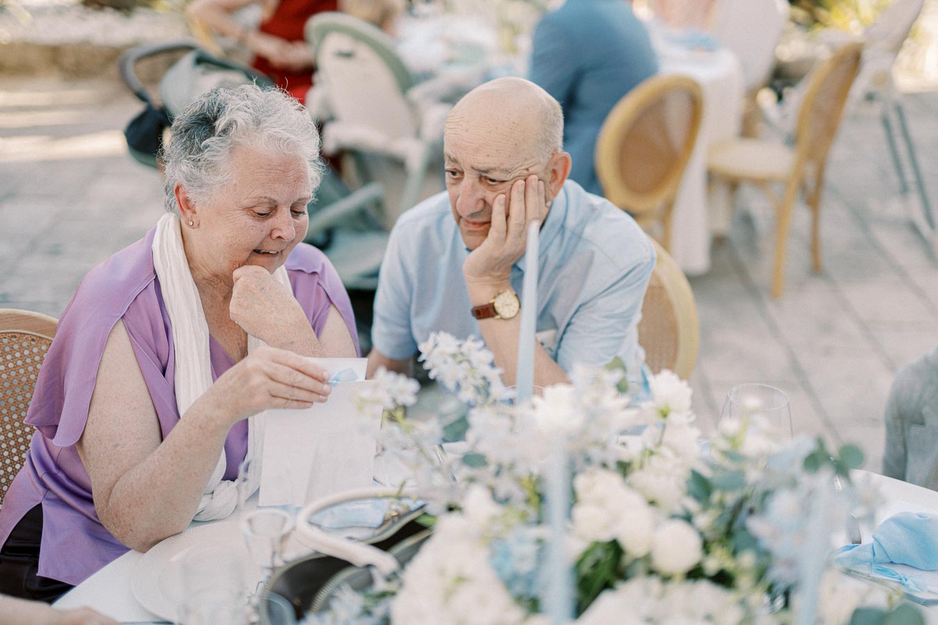 Elderly couple sitting at an outdoor event, reading a card at a table adorned with elegant floral centerpieces.