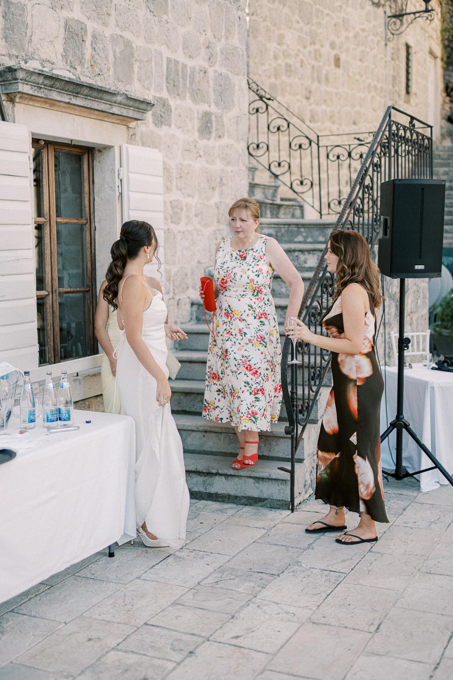 Three women conversing near stone steps at an outdoor event, dressed in elegant summer dresses, with a table set with water bottles in the foreground and wrought iron railings in the background.