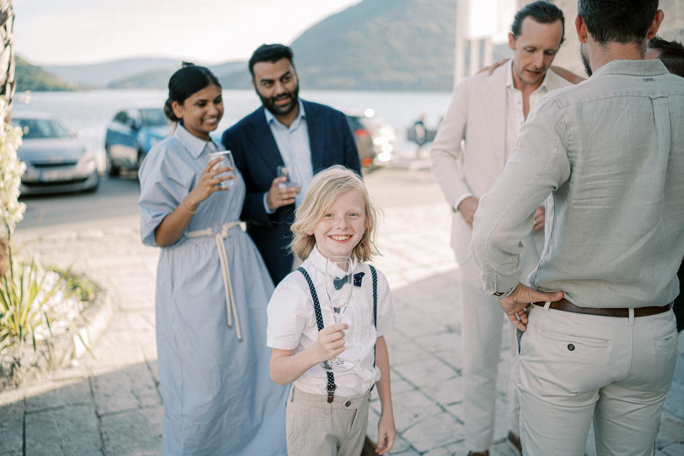 A joyful group of well-dressed people, including a child in suspenders, socializing outdoors near a scenic waterfront with mountains in the background.