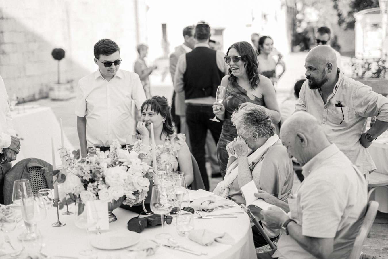 A group of people enjoying a social gathering at an outdoor event, with festive table decorations and floral arrangements in black and white.