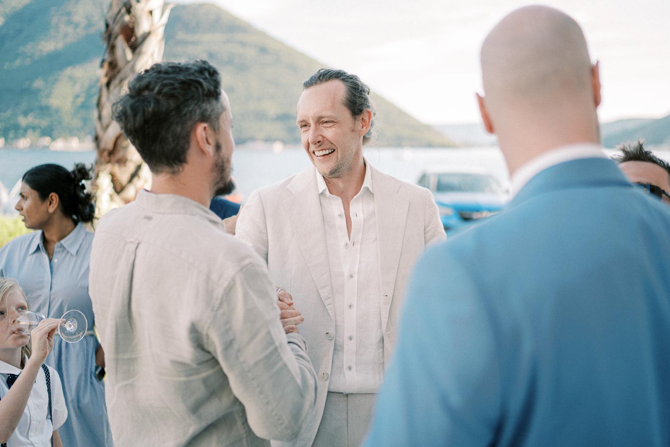 A group of people socializing outdoors at a sunny event near a waterfront, with a mountainous landscape in the background. One person in a light suit is smiling and shaking hands with another, while a child drinks from a glass.