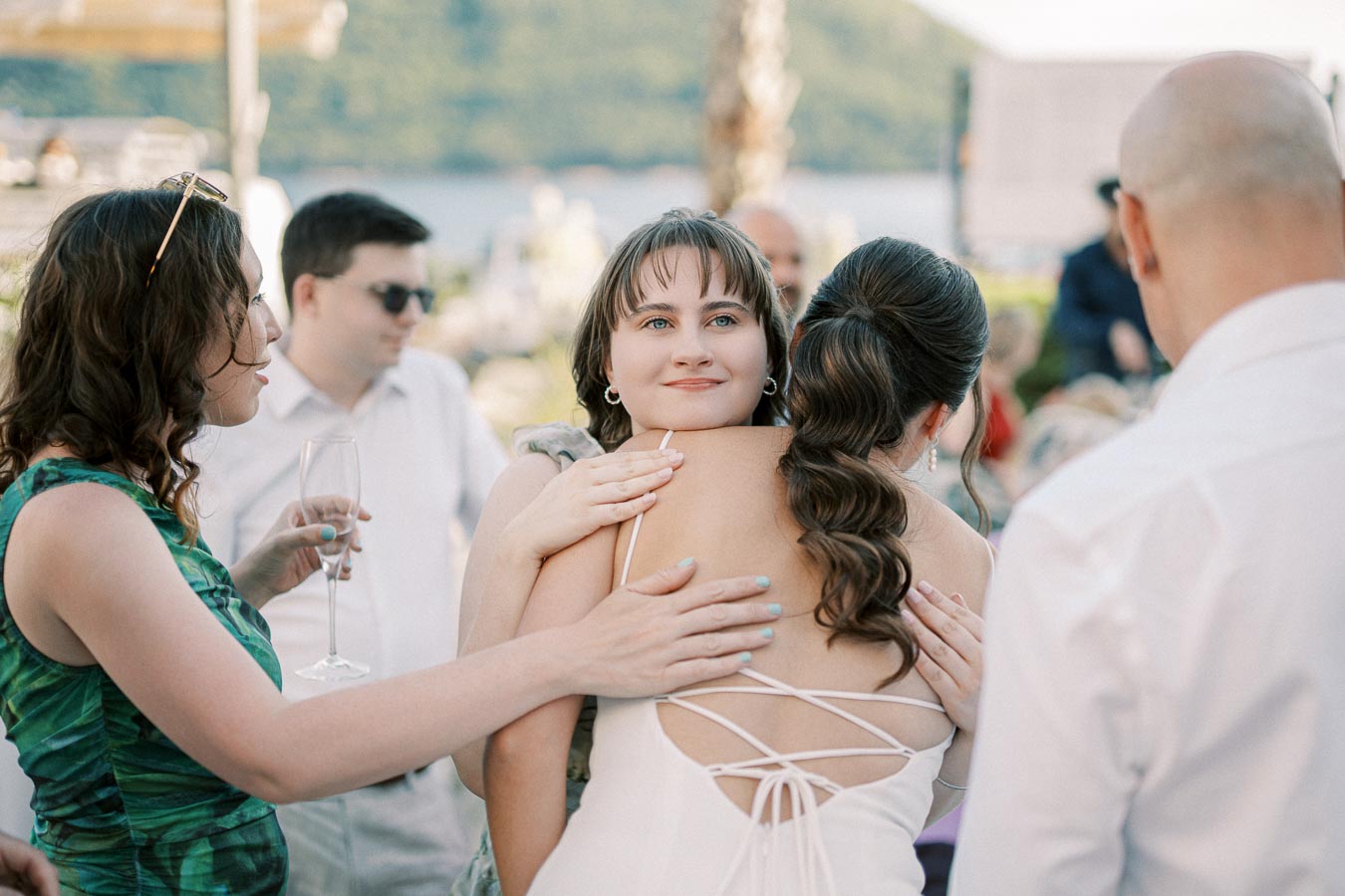 A group of people at an outdoor gathering, with two women embracing warmly, one wearing a white dress with lace-up back. The scene includes a scenic background of lush greenery and a distant body of water, creating a serene and festive atmosphere.