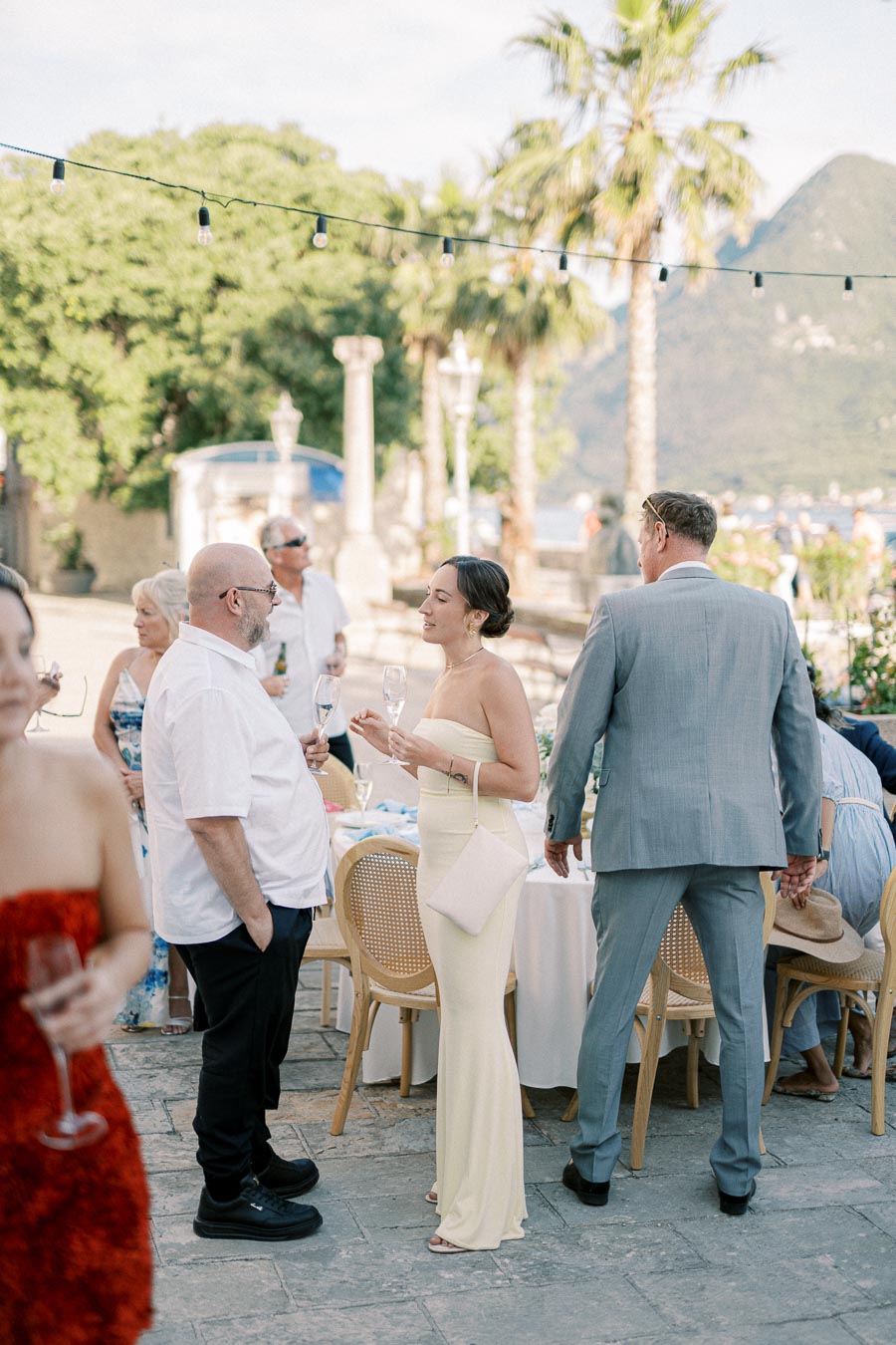 Outdoor social gathering with elegantly dressed guests interacting, surrounded by scenic greenery and mountains, under string lights.