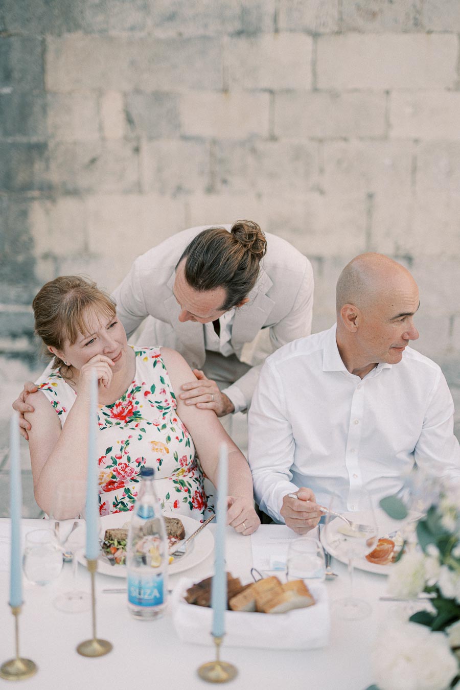 Three people sitting at a beautifully set outdoor table, with one person comforting another who appears emotional. There are candles and a bottle on the table, creating a warm, intimate atmosphere at a gathering.