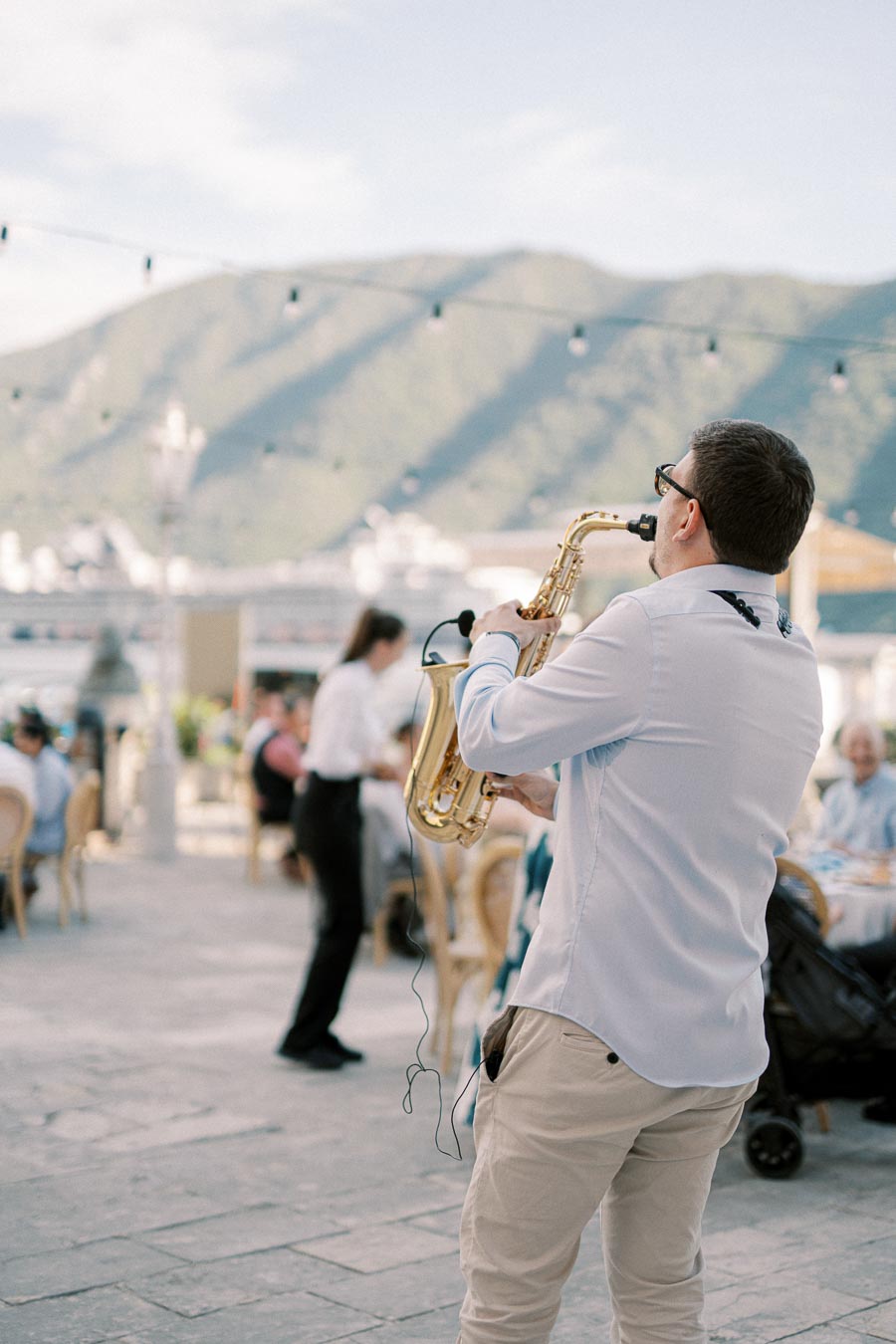 A man in a white shirt plays a saxophone outdoors, with a scenic mountain landscape in the background and people seated at tables, enjoying the music.