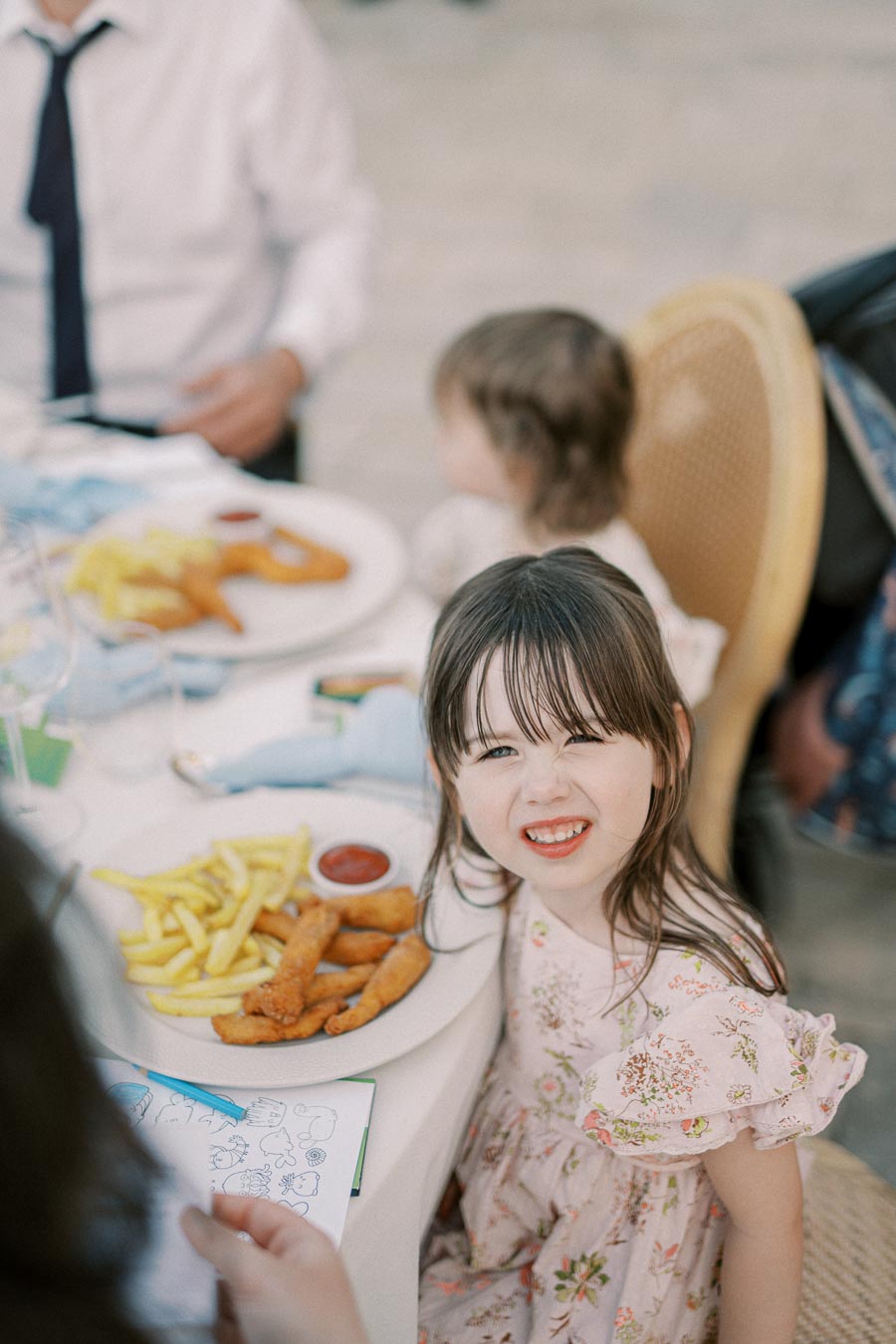 Young girl smiling at a table with a plate of fries and chicken tenders, seated outdoors with family members, creating a joyful family dining experience.
