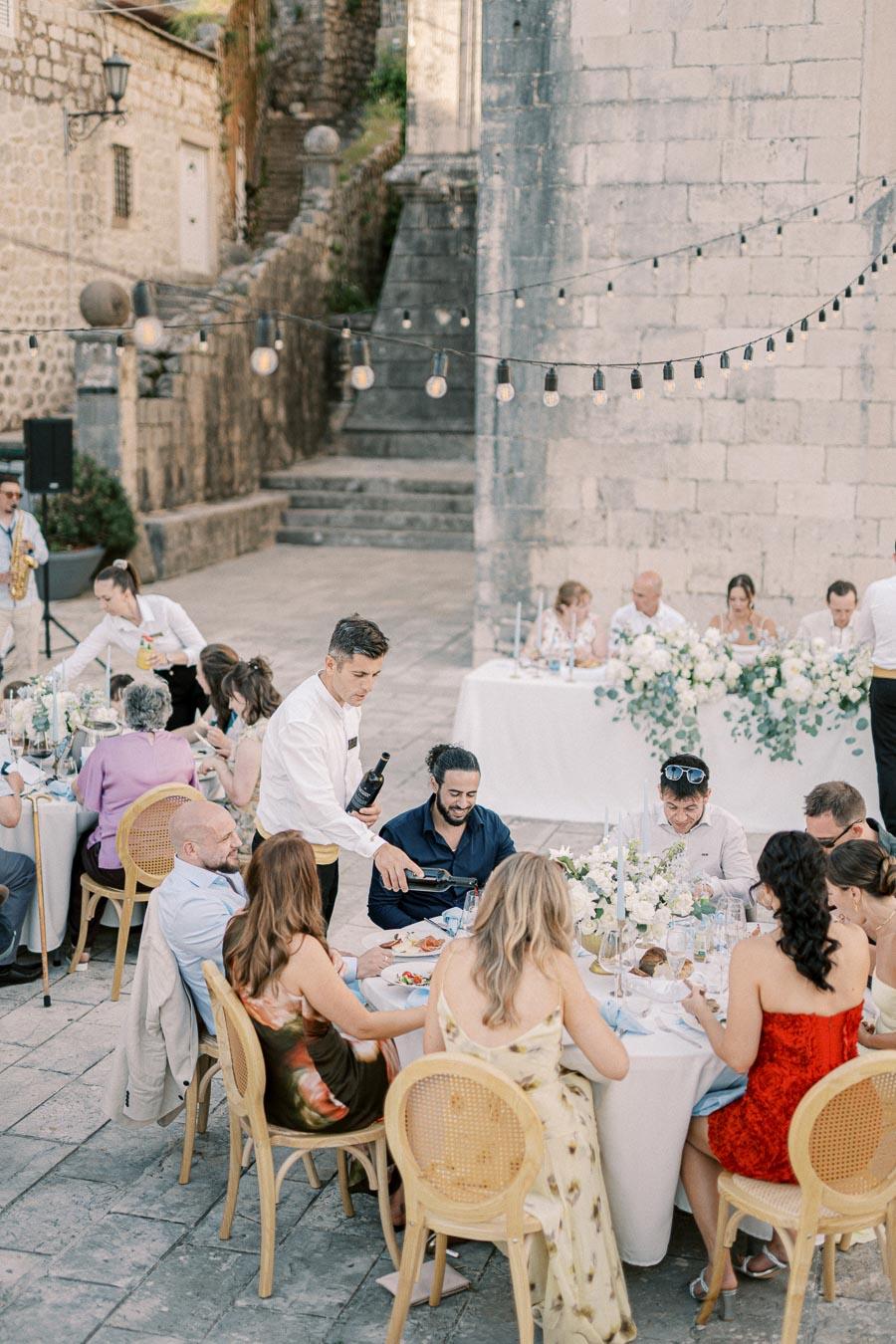 Outdoor wedding reception with guests seated at elegantly decorated tables, enjoying food and drinks under string lights next to a historic stone building.