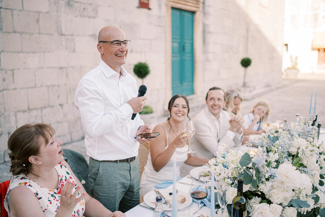 A man in a white shirt giving a speech at an elegant wedding reception, with guests smiling and sitting at a long table adorned with white and blue floral arrangements.