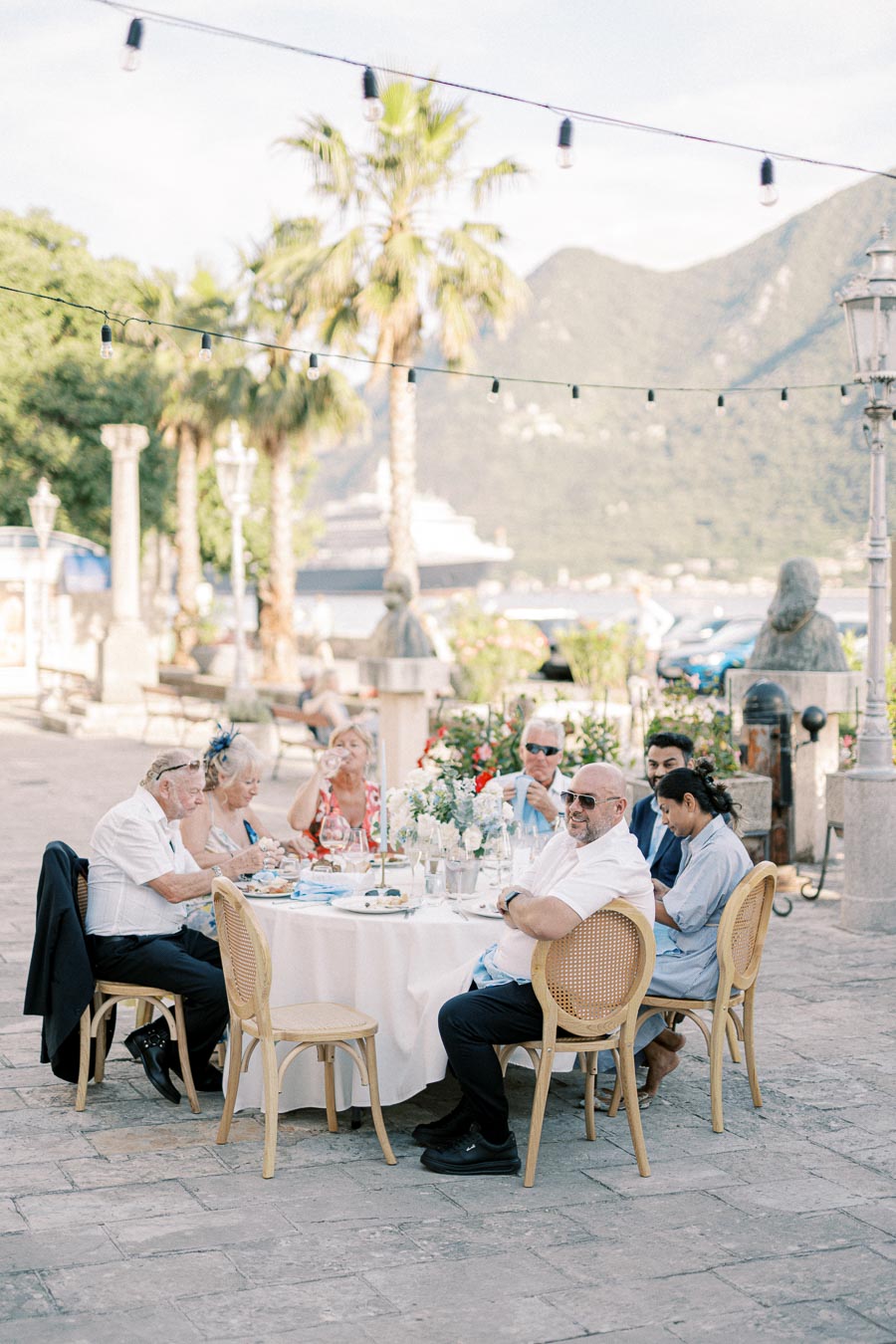 Outdoor gathering at a scenic restaurant with a group of people dining around a table, set against a backdrop of palm trees, picturesque mountains, and a cruise ship in the distance.