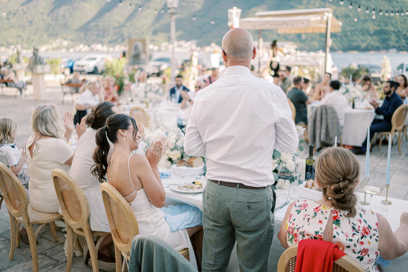Outdoor wedding reception with guests seated at decorated tables, enjoying a scenic view of mountains and water. A man in a white shirt stands giving a speech, while attendees applaud and celebrate.