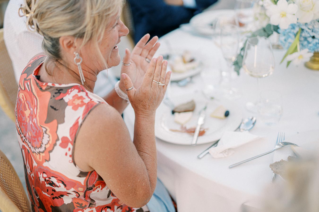 A woman in a floral dress clapping joyfully at an elegantly set white table during an outdoor event, accompanied by assorted appetizers and floral arrangements.
