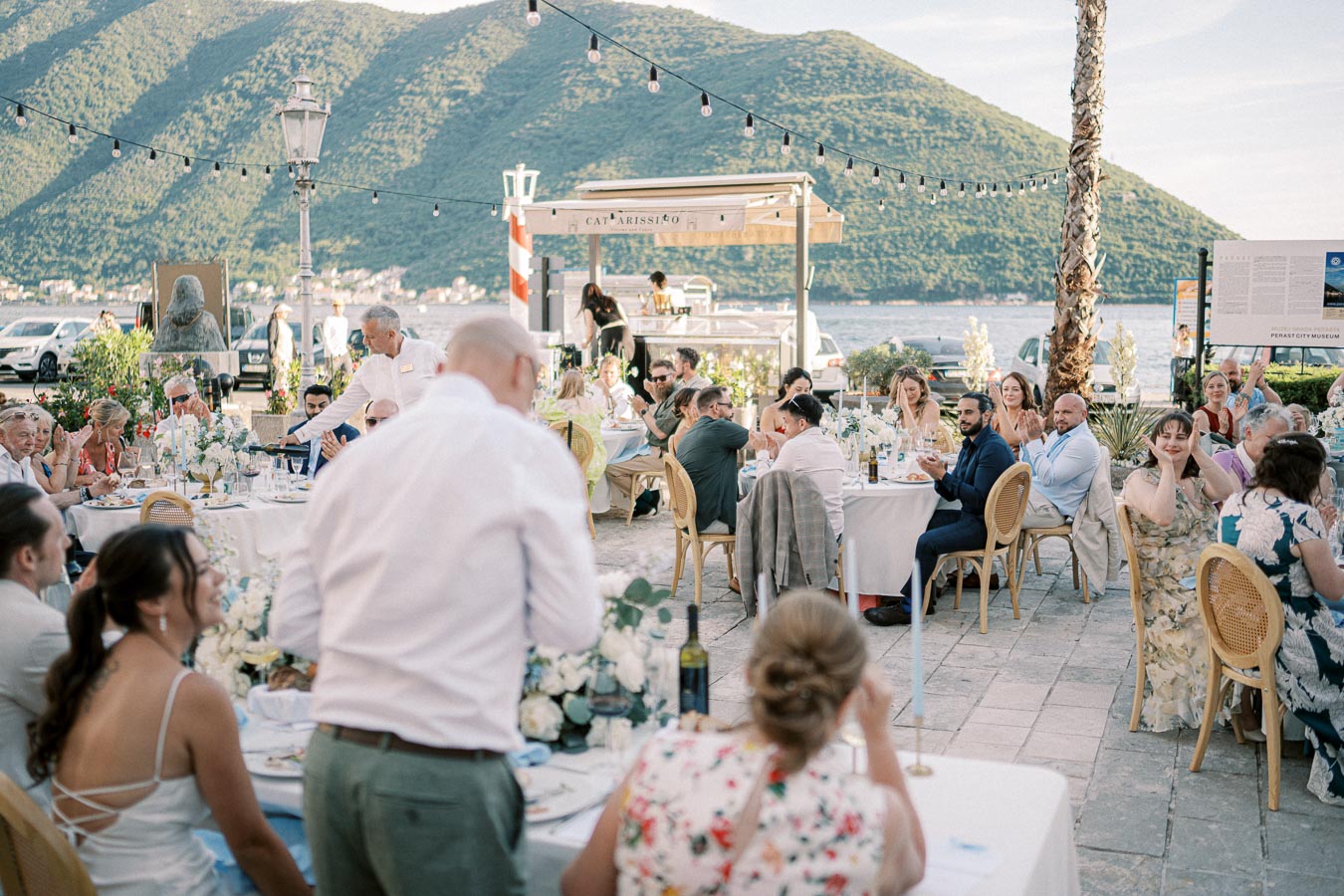 Outdoor wedding reception by the sea, with guests seated at tables decorated with floral centerpieces, surrounded by lush green mountains and string lights.