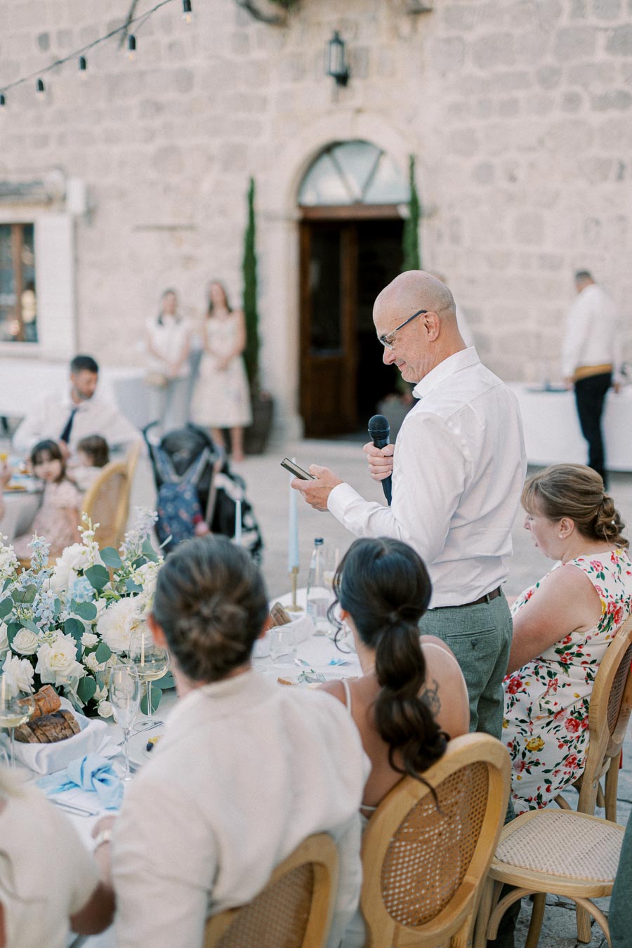 Man giving speech at outdoor wedding reception, holding a microphone and reading notes, surrounded by seated guests and floral decorations.