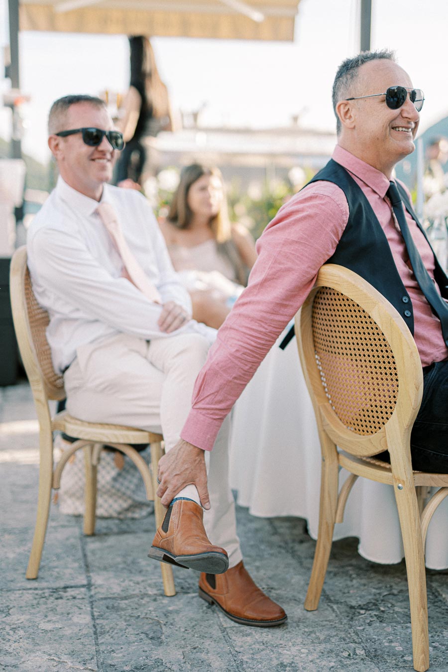 A cheerful man in sunglasses and a red shirt sits on a wooden chair at an outdoor event, holding his ankle above beige shoes on a sunny day.