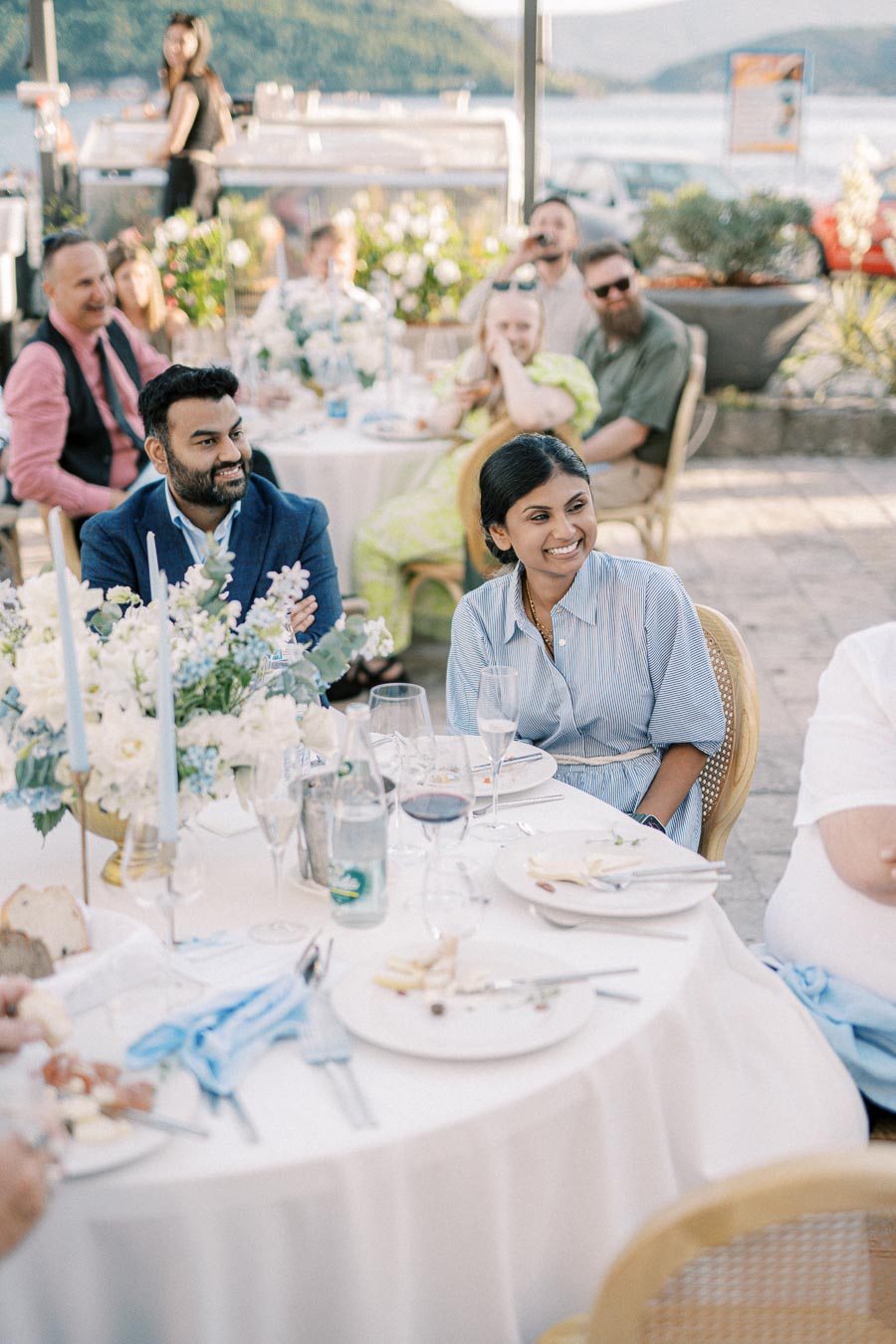 Smiling guests sitting at an elegantly set outdoor table during a sunny garden party, with floral decorations and a scenic background.