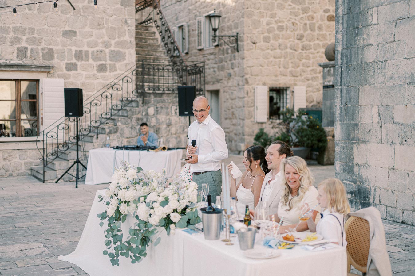 Outdoor wedding reception in a charming stone courtyard, featuring a smiling speaker holding a microphone beside a flower-adorned table with guests enjoying the celebration, while a DJ prepares in the background.