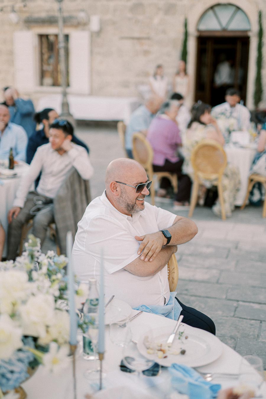 Bald man with sunglasses smiling at an outdoor event with people seated at tables adorned with white and blue decorations.
