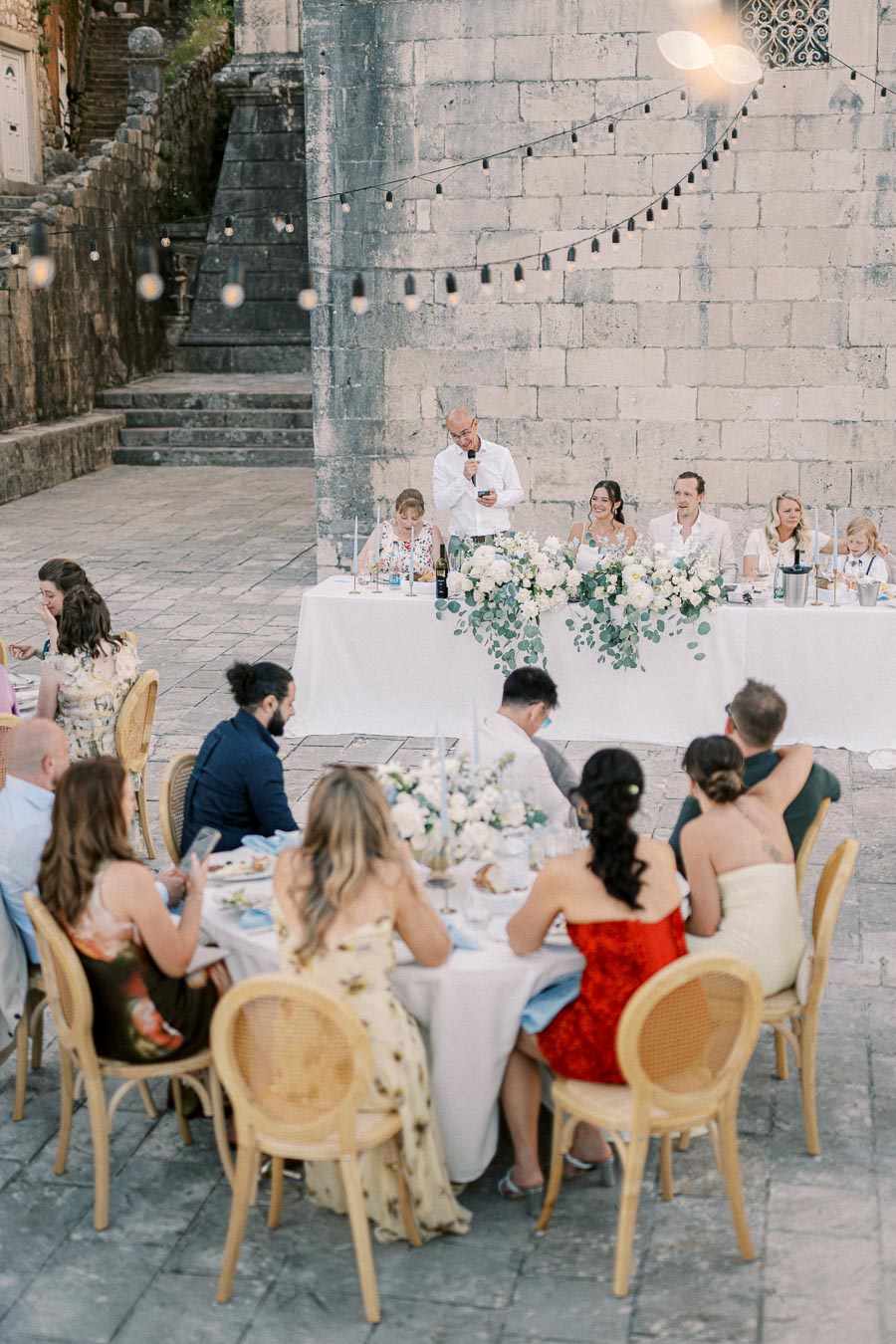 A beautifully decorated outdoor wedding reception with guests seated at round tables, a long head table adorned with white flowers and greenery, people enjoying speeches under string lights against a rustic stone backdrop.