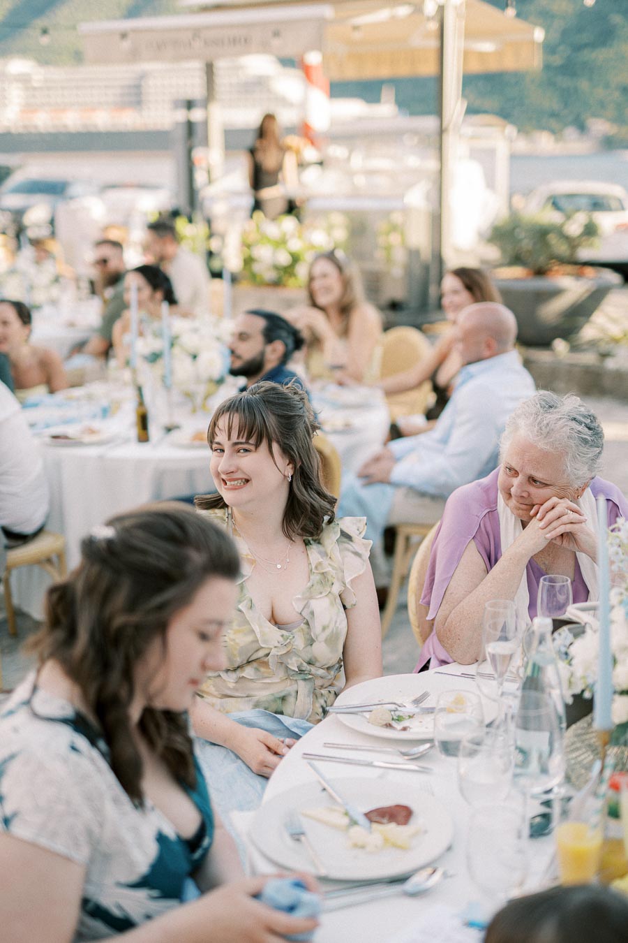 Outdoor wedding reception with guests smiling around decorated tables, featuring floral arrangements and a scenic waterfront backdrop.