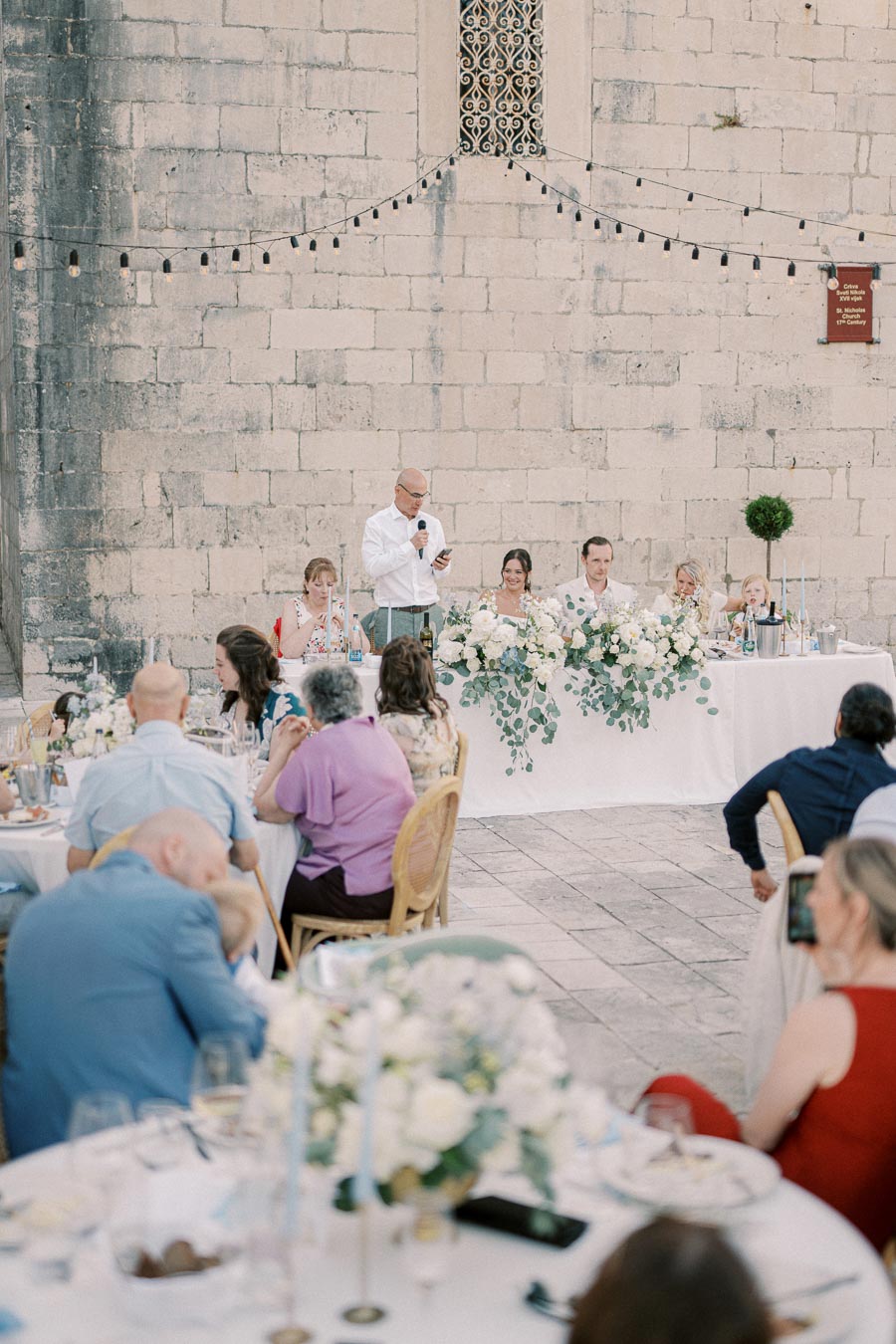 Outdoor wedding reception with guests seated at tables. A man gives a speech at a head table adorned with white flowers and greenery. A historic stone wall and string lights serve as background decor, creating a romantic atmosphere.