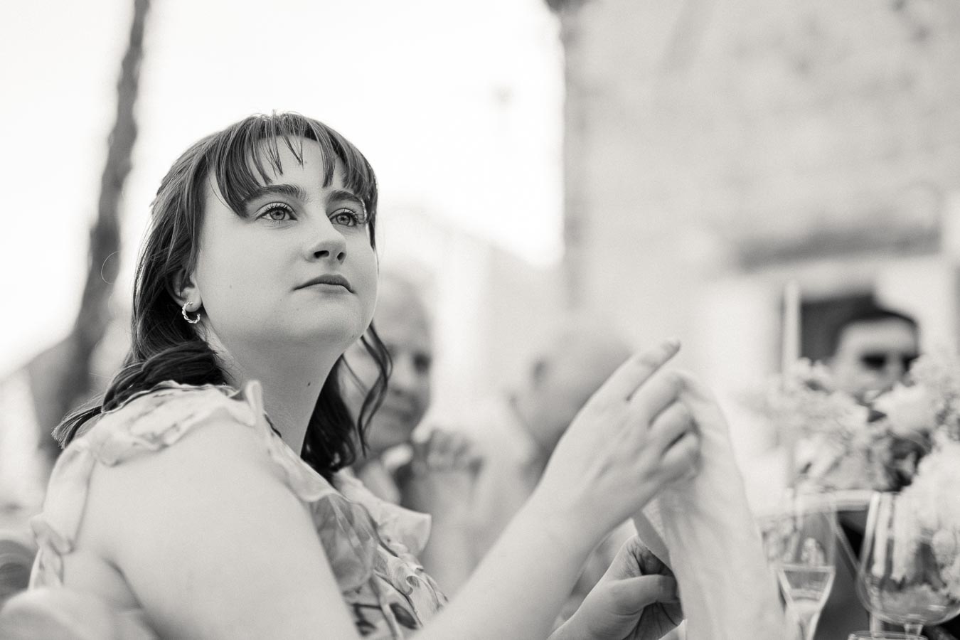 A woman at an outdoor event, seated at a table with a floral centerpiece and glassware, looking thoughtfully into the distance, captured in black and white photography.