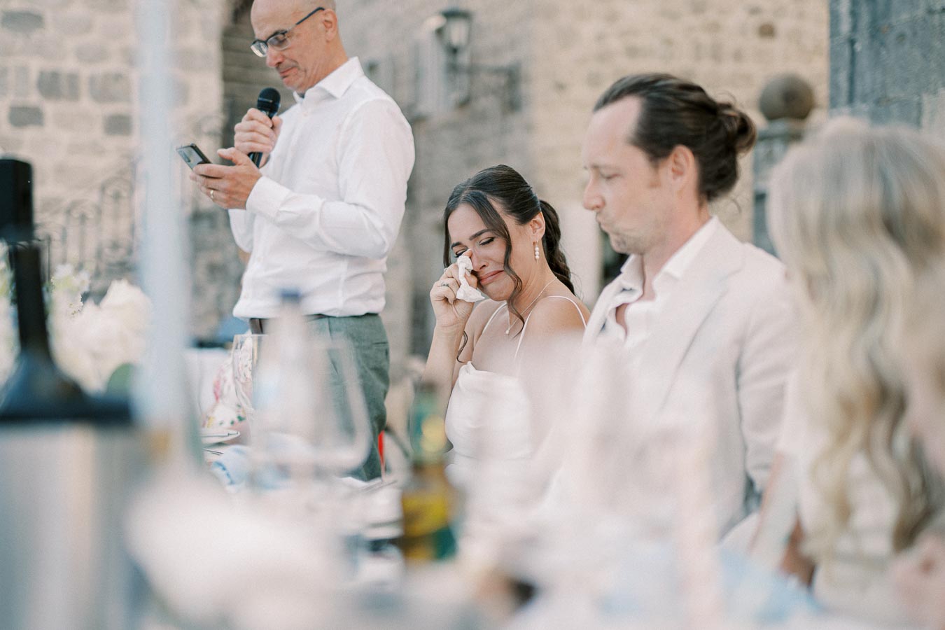 A woman wiping tears during a heartfelt wedding speech, with guests seated nearby at an outdoor reception.