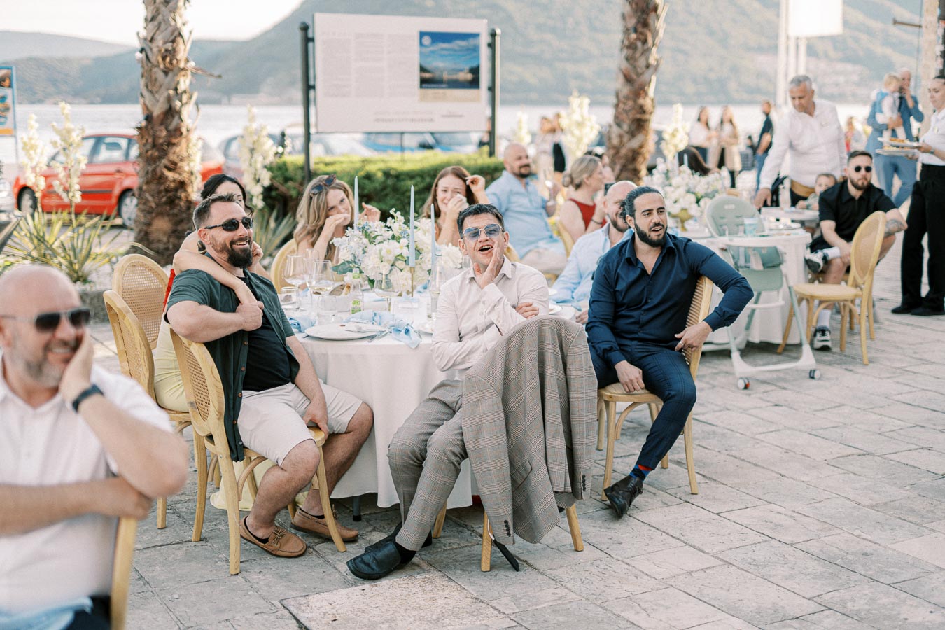 Group of people enjoying a lively outdoor event at a scenic location with mountains and a lake in the background, featuring elegantly set round tables and palm trees lining the venue.