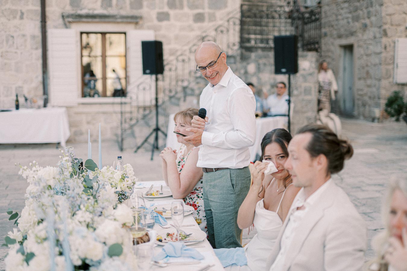 A man in a white shirt and glasses gives a speech at an outdoor wedding reception, holding a microphone. Guests are seated at a decorated table with floral arrangements and candles, with a woman wiping her tears. Stone building and speakers in the background.