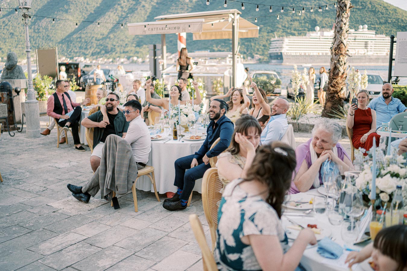 Outdoor wedding reception with guests seated at decorated tables, enjoying the event under string lights. Scenic mountain and sea views in the background, creating a festive and picturesque atmosphere.