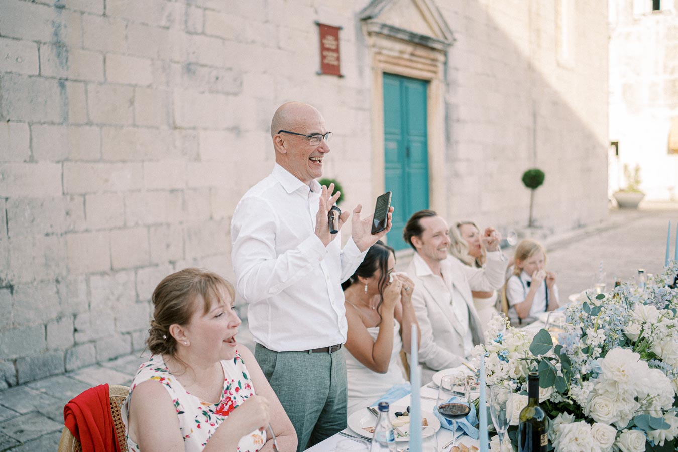 A joyful man giving a speech at an outdoor wedding reception, surrounded by smiling guests seated at a beautifully decorated table with elegant white and blue floral arrangements, set against a rustic stone building backdrop.