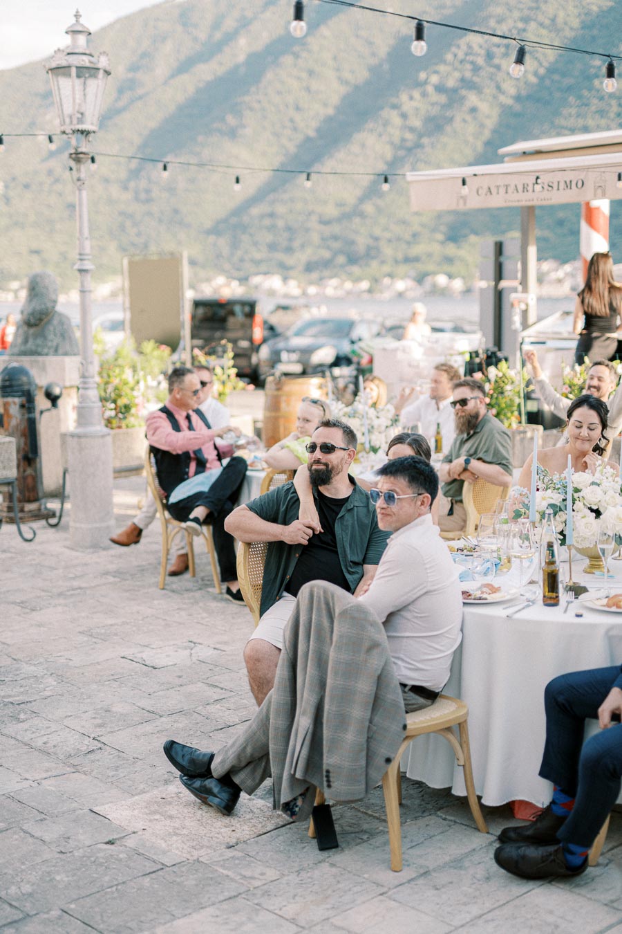 A lively outdoor dining scene with people seated at tables decorated with flowers, set against a backdrop of mountains and string lights under a clear sky.