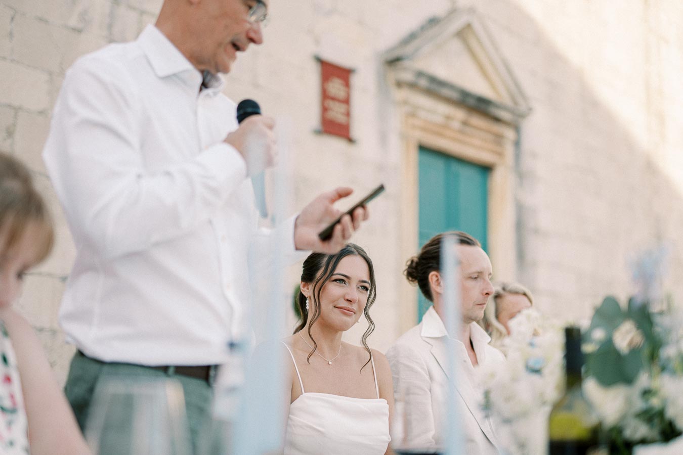 A man gives a speech at a wedding reception, standing beside a smiling woman in a white dress, with another man seated nearby in a light suit, surrounded by flowers and decorative elements.
