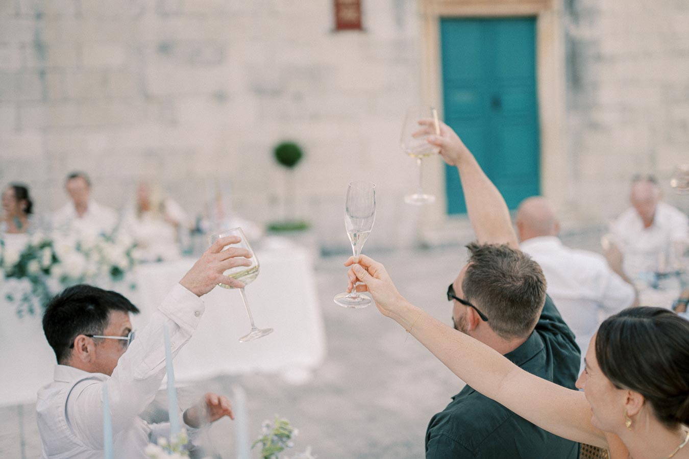 Outdoor wedding celebration with guests raising glasses in a toast, featuring a formal setting with a white tablecloth and floral centerpieces in front of a stone building.