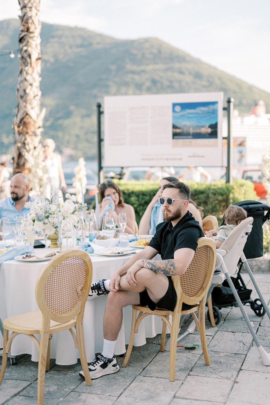 Outdoor dining event with people seated around a table, featuring elegant decor and a scenic mountain backdrop. Casual summer attire with a relaxed atmosphere.