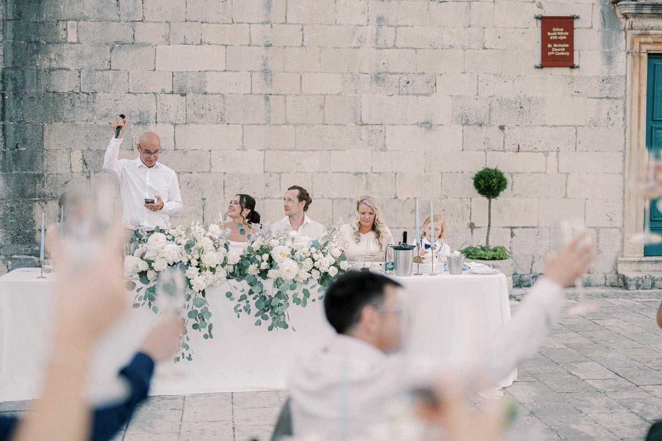 A wedding reception scene with a bride and groom seated at a decorated table, surrounded by guests raising glasses. The background features an old stone wall with a plaque indicating St. Nicholas Church, 13th century. The atmosphere is festive and elegant, with white floral arrangements enhancing the elegant setting.
