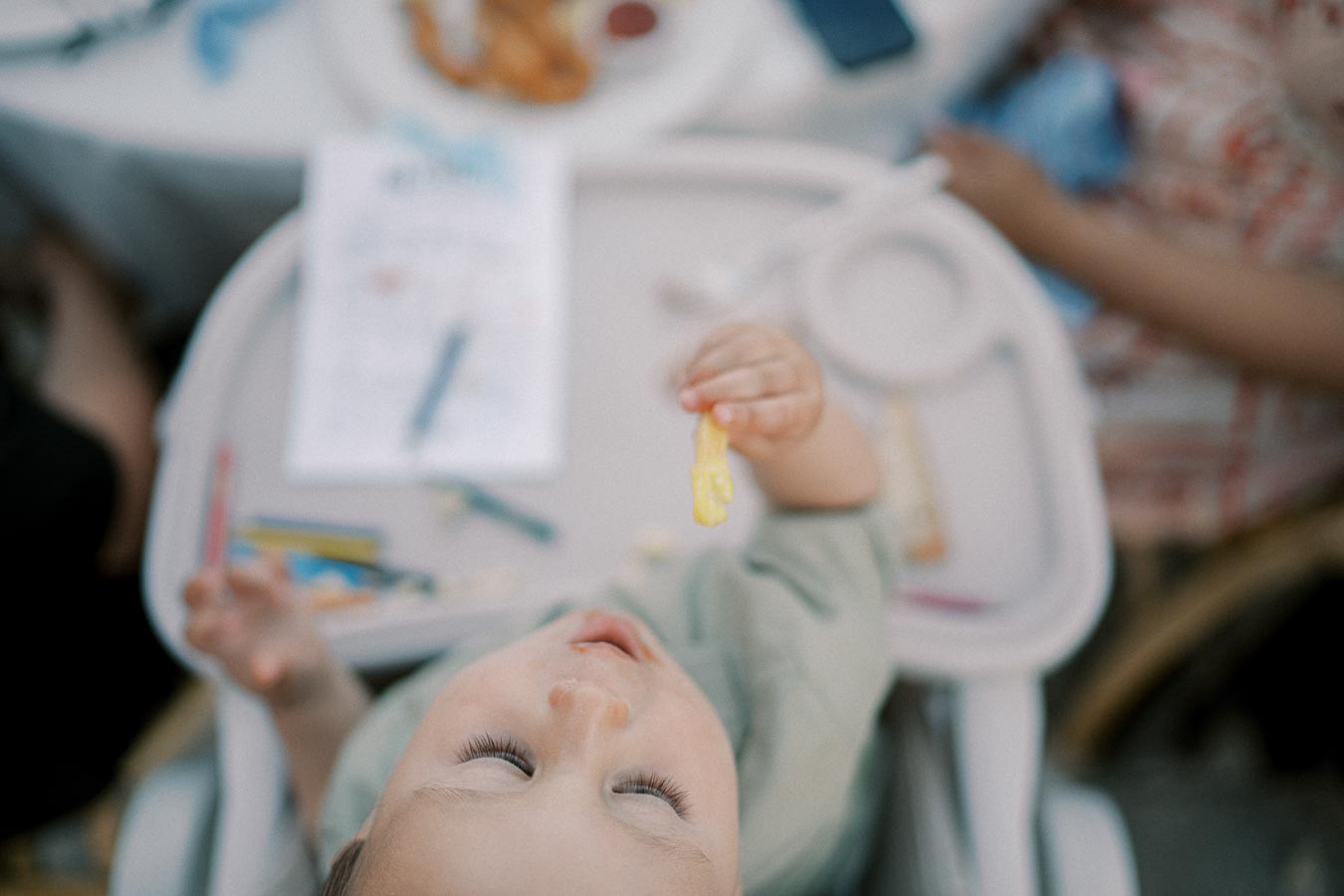 A toddler sitting in a high chair, holding a piece of yellow food, surrounded by crayons and paper with food on the table in the background.