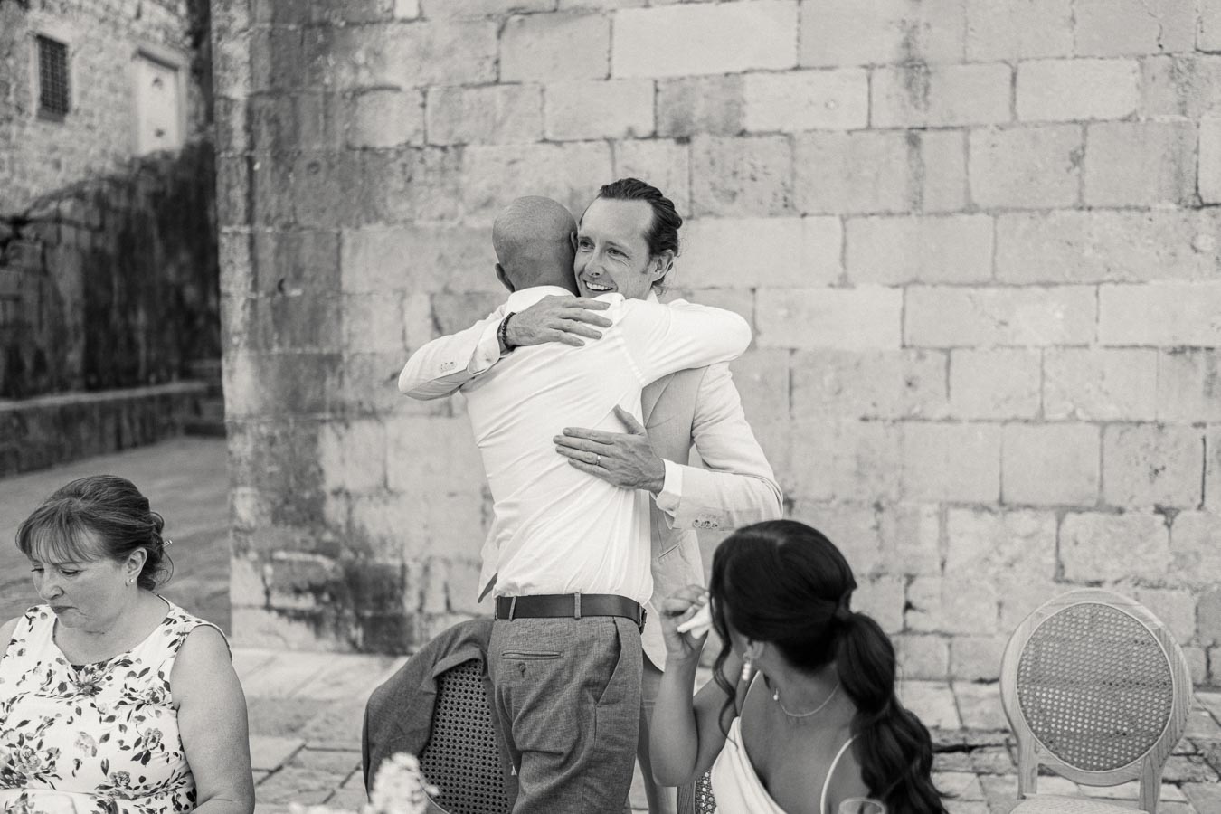 Black and white photo capturing an emotional moment at an outdoor event. Two men embrace warmly, celebrating friendship against a stone wall backdrop. A woman in floral attire sits at a table, and another woman wipes tears, adding to the heartfelt atmosphere.