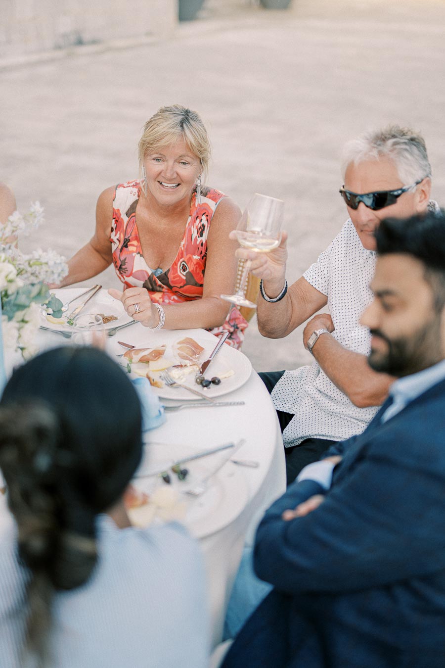 A group of people enjoying an outdoor meal, with a woman in a floral dress smiling while a man holds a glass of wine, sitting at a round table with plates of food.