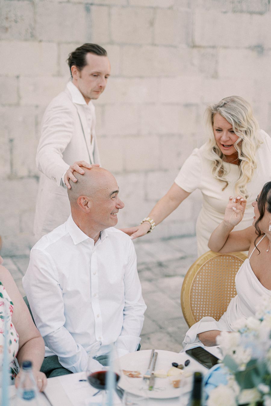Group of people enjoying a lively conversation during an outdoor social gathering, with a man in a white shirt smiling as another man playfully places a hand on his head, surrounded by a woman reaching out, and a table set with dishes and drinks.
