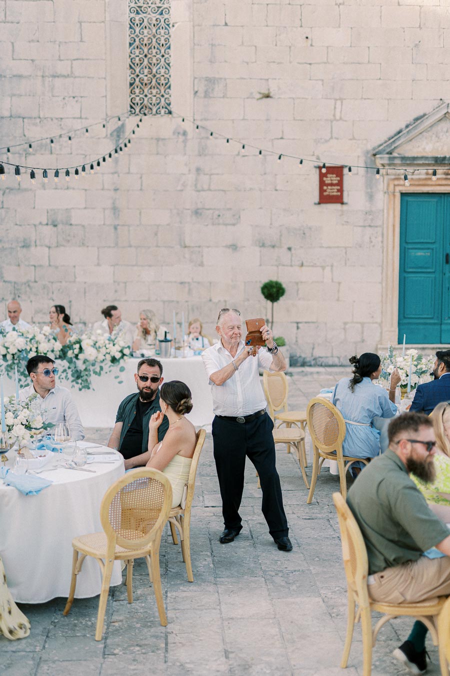 Outdoor wedding reception in a European courtyard, featuring guests seated at elegantly decorated tables with white floral arrangements. A man in a white shirt is capturing a photo with a vintage camera, while others enjoy the celebration under string lights.
