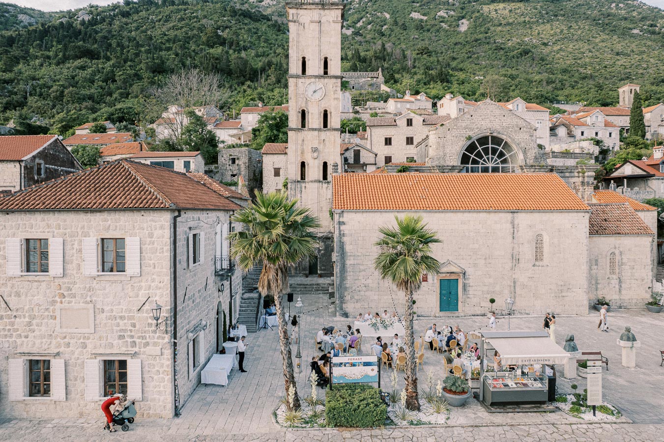 Aerial view of a charming European village square in Perast, Montenegro, featuring historic stone buildings with red-tiled roofs. A bell tower rises in the center, surrounded by lush green hills. Tables with dining people and palm trees enhance the picturesque setting.