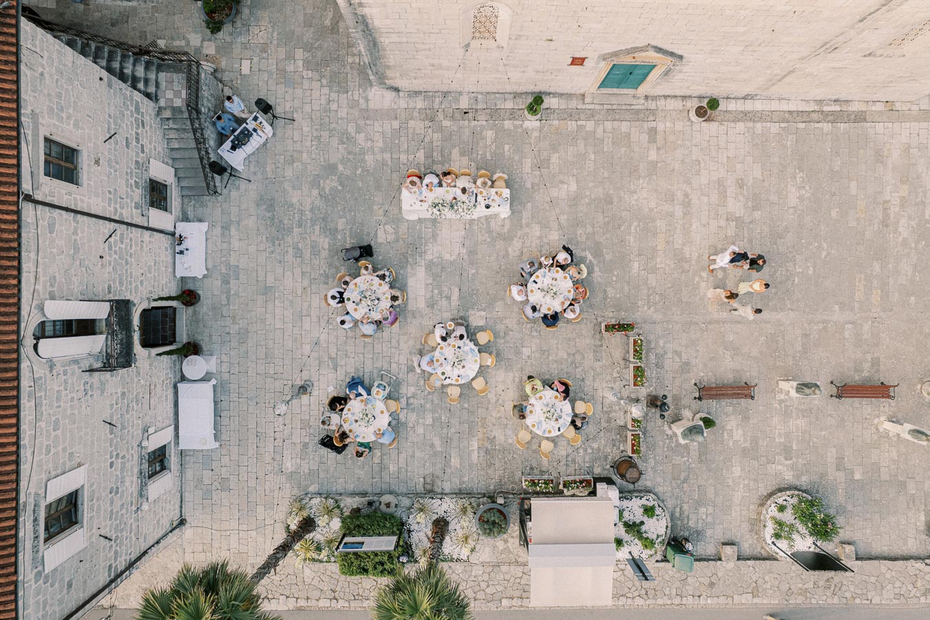 Aerial view of an outdoor wedding reception with round tables set up for guests in a stone courtyard. Guests are seated and socializing, surrounded by historic architecture, flowers, and greenery.