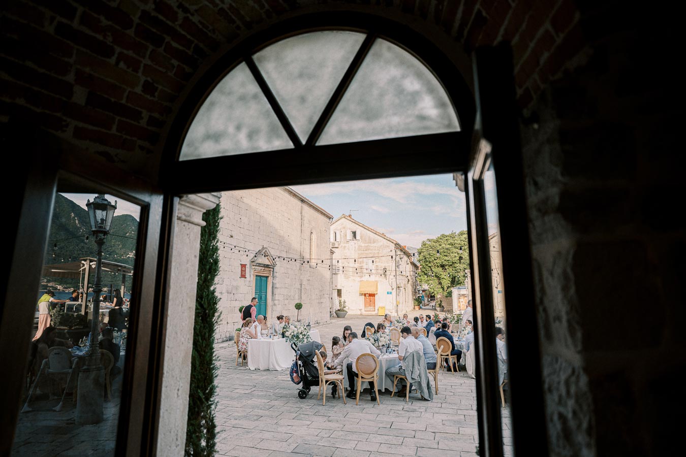 Outdoor wedding reception scene viewed through an arched doorway, featuring guests seated at elegantly decorated tables in a historic stone courtyard setting.
